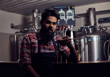 Şık tam Hintli bir yün gömlekli sakallı ve önlük bir bardak bira, bira fabrikası sayacında arkasında oturan tutar.