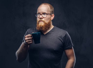 Kızıl saçlı gözlüklü karanlık dokulu bir arka plan üzerinde izole bir gri t-shirt giymiş kahve ile erkek ayakta sakallı.