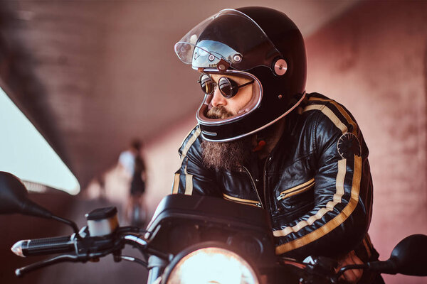 Close-up portrait of a brutal bearded biker in helmet and sunglasses dressed in a black leather jacket sitting on a retro motorcycle with included headlight.