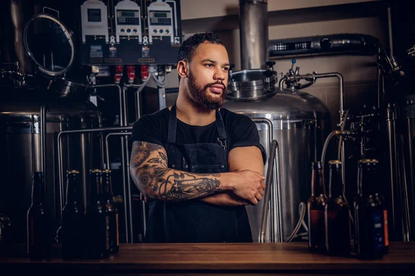 Portrait of a stylish bearded tattooed dark skinned male with crossed arms standing behind the counter in the brewery.
