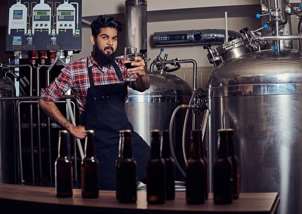 时髦的满胡子的印度男人在一个羊毛衬衫和围裙持有一杯啤酒 站在柜台后面的啤酒厂 — 图库照片