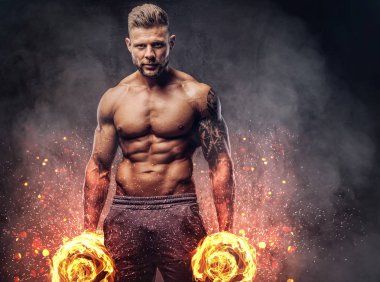 Kendinden emin bir bakışla kameraya bakarak bir stüdyoda dumbbells ile poz güçlü şık vücut geliştirmeci, kolundaki dövme ile. Yangın sanat kavramı.
