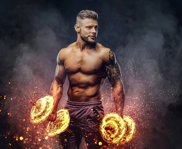 Powerful stylish bodybuilder with tattoo on his arm, doing the exercises with dumbbells. Fire art concept.