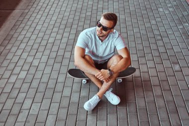 Beyaz bir gömlek ve şort bir kaykay uzağa arıyorsunuz bir patika üzerinde oturan güneş gözlüklü bir yakışıklı şık patenci adam portresi üstten görünüm giymiş.