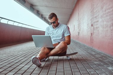 Beyaz bir gömlek ve şort köprü altında bir kaykay üzerinde otururken bir dizüstü bilgisayarda çalışma portre yakışıklı bir moda Freelancer güneş gözlüğü giymiş.