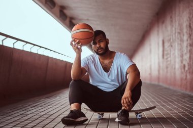 Dalgın bir esmer adam portresi beyaz bir gömlek giymiş ve spor şort tutan bir basketbol footway köprü altında bir kaykay üzerinde otururken.