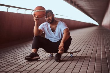 Dalgın bir esmer adam portresi beyaz bir gömlek giymiş ve spor şort tutan bir basketbol footway köprü altında bir kaykay üzerinde otururken.