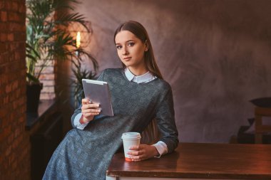 Genç kız zarif bir gri elbise giymiş bir paket servisi olan restoran kahve tutar ve bir tablet tutan bir oda çatı iç tablo yaslanmış, bir kameraya bakıyor.