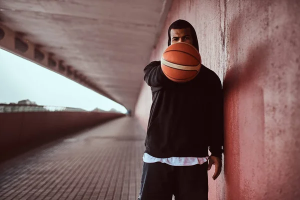Portrait of a dark-skinned guy dressed in a black hoodie and sports shorts holds a basketball while standing on a footway under a bridge.