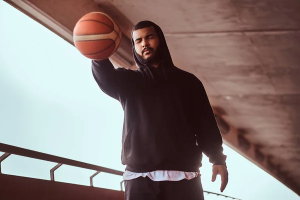 Portrait of a dark-skinned guy dressed in a black hoodie and sports shorts holds a basketball while standing on a footway under a bridge.