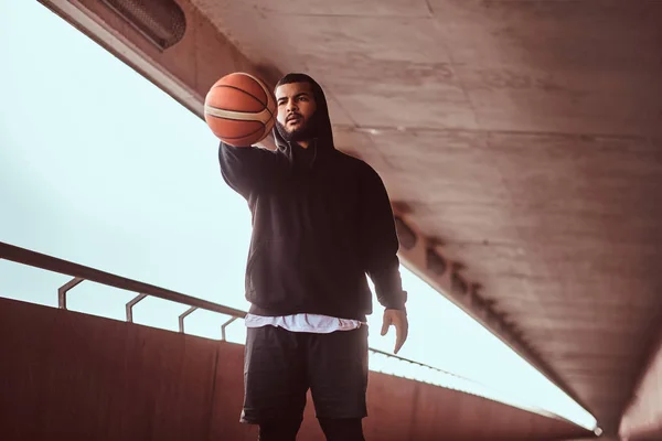 Portrait of a dark-skinned guy dressed in a black hoodie and sports shorts holds a basketball while standing on a footway under a bridge.