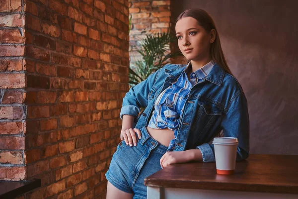 一个年轻的时髦女孩的肖像穿着衬衫和牛仔裤夹克和短裤倚在桌子上与一杯外卖咖啡在一个房间内阁楼内部 — 图库照片