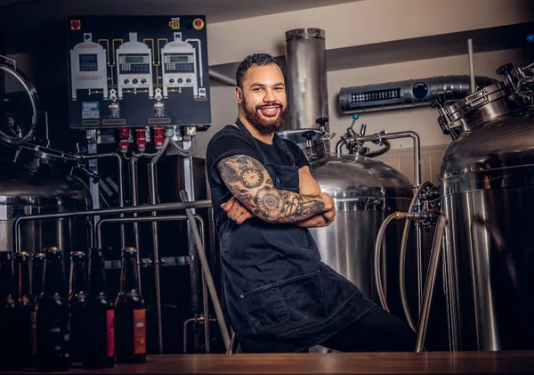 Happy stylish bearded tattooed dark-skinned male with crossed arms standing behind the counter in a brewery.