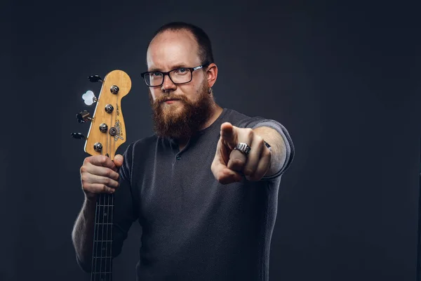 Redhead bearded male musician wearing glasses dressed in a gray t-shirt holds electric guitar. Isolated on dark textured background.