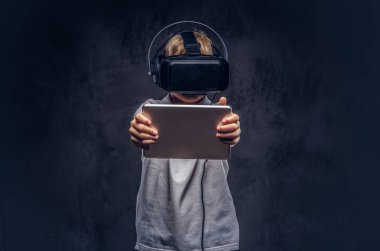 Sarı saçlı Genç çocuk sanal gerçeklik gözlük takan ve dijital tablet kullanma beyaz bir t-shirt giymiş. Karanlık dokulu bir arka plan üzerinde izole.