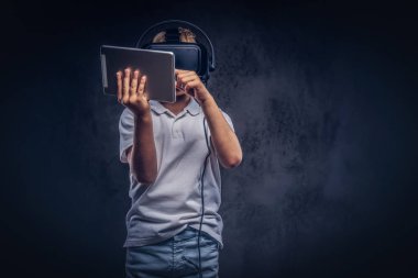 Liseli Sarışın sanal gerçeklik gözlük takan ve dijital tablet kullanma beyaz bir t-shirt giymiş. Karanlık dokulu bir arka plan üzerinde izole.