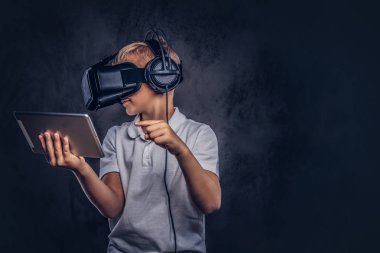 Liseli Sarışın sanal gerçeklik gözlük takan ve dijital tablet kullanma beyaz bir t-shirt giymiş. Karanlık dokulu bir arka plan üzerinde izole.