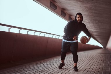Adam koyu tenli bir footway köprü altında basketbol oynamak siyah bir hoodie ve spor şort giymiş.