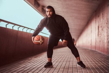 Bir kameraya bakarak adam koyu tenli bir footway bir köprü altında basketbol oynamak siyah bir hoodie ve spor şort giymiş.