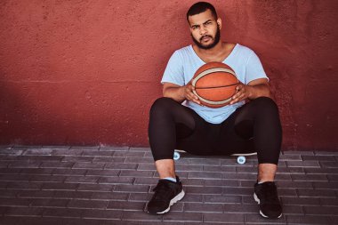 Koyu renkli sakallı adam portresi basketbol ile bir kaykay üzerinde oturan ve bir duvara yaslanmış bir beyaz gömlek ve spor şort giymiş.
