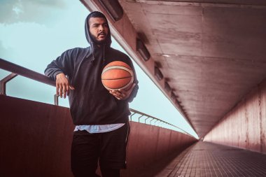 Uzağa arıyorsunuz sakallı adam koyu tenli bir Otokorkuluk duran ile basketbol, yaslanmış bir siyah hoodie ve spor şort giymiş.