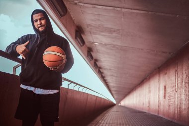 Serin koyu renkli bir Otokorkuluk eğilerek basketbol ile ayakta, bir kameraya bakarak siyah bir hoodie ve spor şort giymiş adam sakallı.