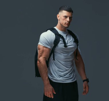 Kas yakışıklı vücut geliştirmeci spor sırt çantası gri bir arka planda poz ile.