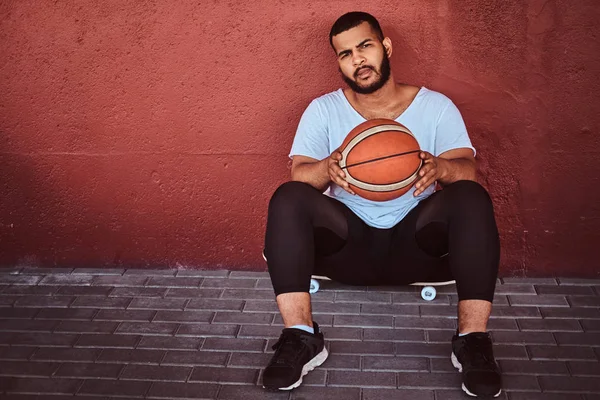 Koyu renkli sakallı adam portresi basketbol ile bir kaykay üzerinde oturan ve bir duvara yaslanmış bir beyaz gömlek ve spor şort giymiş.