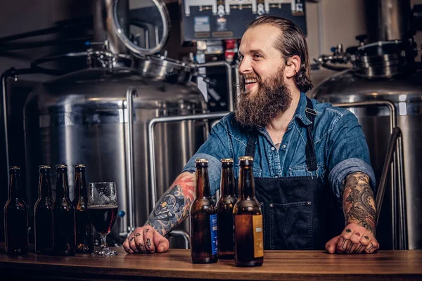 Portrait of a happy bearded tattooed hipster male in a jeans shirt and apron working in a brewery factory, standing behind the counter.
