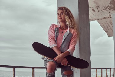 Sarı saçlı mutlu çekici genç kız portresi pembe bir ceket giymiş ve kot tutan bir Otokorkuluk siyah beyaz deniz kıyısında arka plan üzerinde otururken bir kaykay sökük.