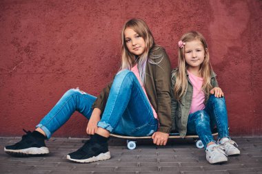 İki küçük kız portresi Köprüsü footway adlı bir kaykay üzerinde otururken bir duvara yaslanmış trendy kıyafetler giymiş.