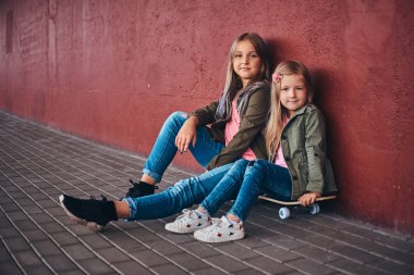İki sevimli küçük kız portresi Köprüsü footway adlı bir kaykay üzerinde otururken bir duvara yaslanmış trendy kıyafetler giymiş.