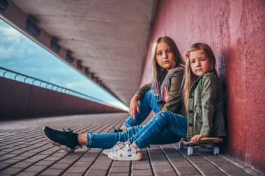 İki küçük kız portresi Köprüsü footway adlı bir kaykay üzerinde otururken bir duvara yaslanmış trendy kıyafetler giymiş.