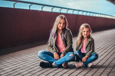İki küçük kız portresi birlikte bir köprü footway adlı bir kaykay üzerinde oturan trendy kıyafetler giymiş.