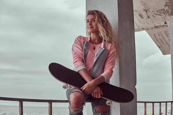 一个快乐迷人的年轻女孩的肖像身着粉红色夹克和撕裂牛仔裤举行滑板 而坐在一个护栏上的黑色和白色的海海岸背景 — 图库照片