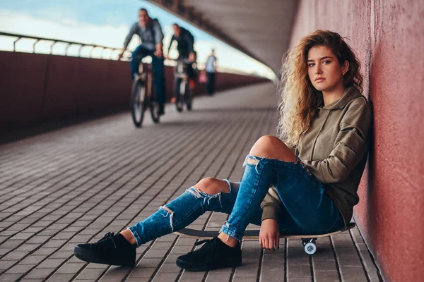 若い女性の肖像画のパーカーを着たし 橋通行でスケート ボードに座って壁にもたれジーンズをリッピング — ストック写真