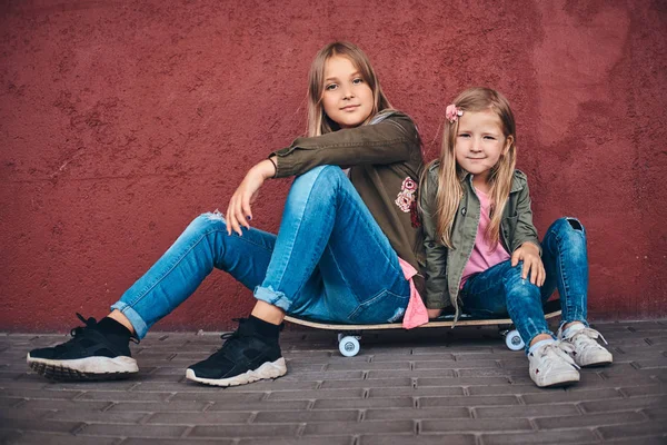 两个妹妹的肖像穿着时髦的衣服倚在墙上 而坐在一个滑板在桥人行道 — 图库照片