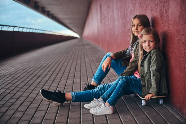 两个妹妹的肖像穿着时髦的衣服倚在墙上 而坐在一个滑板在桥人行道 — 图库照片
