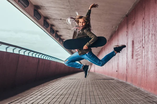 ブロンドの髪の幸せな女子高生は橋通行でスケート ボードでジャンプ トレンディな服を着た — ストック写真