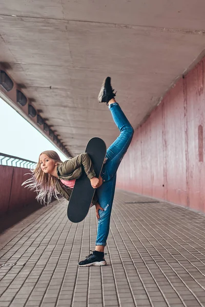 Schoolgirl with blonde hair dressed in trendy clothes dance with a skateboard at a bridge footway.