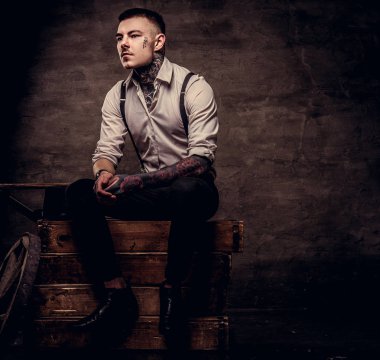 Eski kafalı beyaz gömlek ve pantolon askısı ahşap kutular bir stüdyoda oturup..... giyen adam dövme. Karanlık dokulu bir arka plan üzerinde izole.