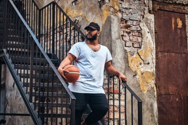 Sakallı bir Afrikalı-Amerikalı portresi beyaz bir t-shirt ve spor şort giymiş şapkalı adam tutan bir Getto merdivenlerde durup, uzağa arıyorsunuz bir basketbol.