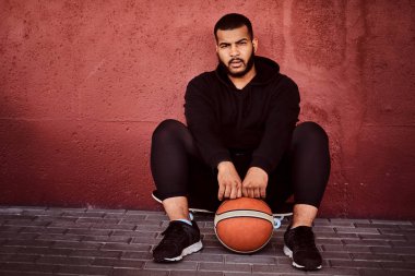 Afrikalı-Amerikalı basketbol ile bir kaykay üzerinde oturan ve duvara yaslanmış bir siyah hoodie ve spor şort giymiş adam sakallı.