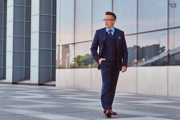 Fiducioso uomo d'affari vestito con un abito elegante distogliendo lo sguardo e tenendo la mano in tasca mentre cammina per strada all'aperto su uno sfondo paesaggio urbano . — Foto Stock