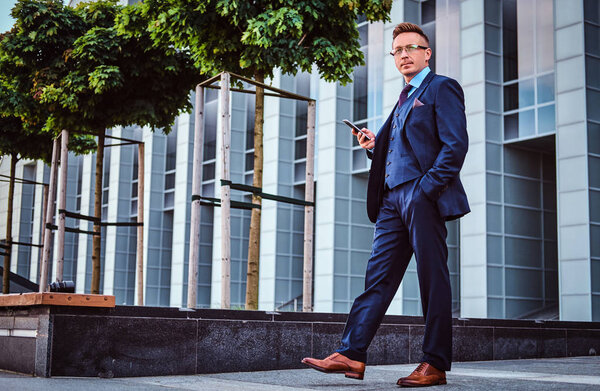 Portrait of a confident stylish businessman dressed in an elegant suit holds a smartphone and looking away while standing outdoors against a skyscraper background.