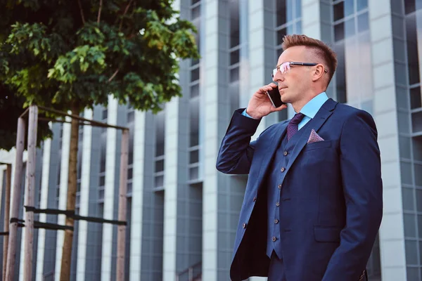 Retrato de un hombre de negocios con estilo y confianza vestido con un traje elegante habla por teléfono y mirando hacia otro lado mientras está parado al aire libre contra el fondo del rascacielos . —  Fotos de Stock