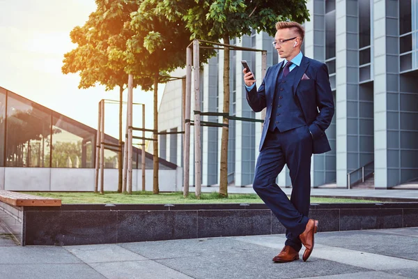 Retrato de un hombre de negocios con estilo y confianza vestido con un traje elegante usando un teléfono inteligente mientras está parado al aire libre contra el fondo del rascacielos . —  Fotos de Stock