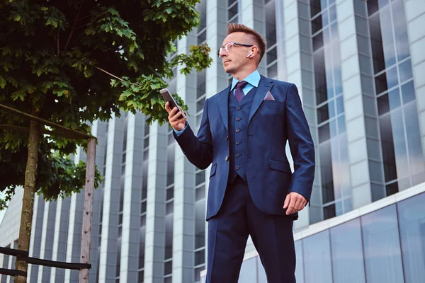 Ritratto di un uomo d'affari elegante e sicuro di sé vestito con un abito elegante tiene uno smartphone e distoglie lo sguardo mentre si trova all'aperto su uno sfondo grattacielo . — Foto Stock