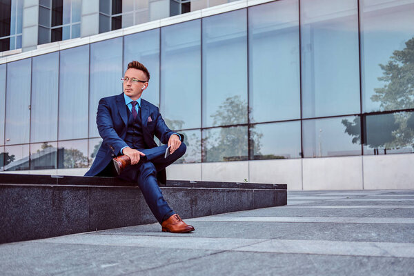 Portrait of a confident stylish businessman dressed in an elegant suit holds a smartphone and looking away while sitting outdoors against a skyscraper background.