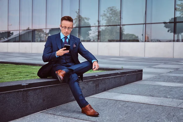 Πορτρέτο ενός αυτοπεποίθηση κομψό επιχειρηματία ντυμένοι με ένα κομψό κοστούμι χρησιμοποιώντας ένα smartphone, ενώ κάθεται σε εξωτερικούς χώρους ουρανοξύστης φόντο. — Φωτογραφία Αρχείου